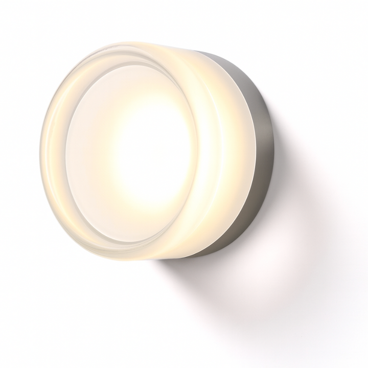 Transparent glass round wall sconce glowing warm white beside a bedside table