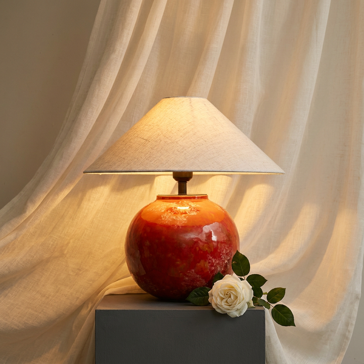 Embrale earth-red ceramic table lamp with cream fabric shade against sheer linen veil, hero shot